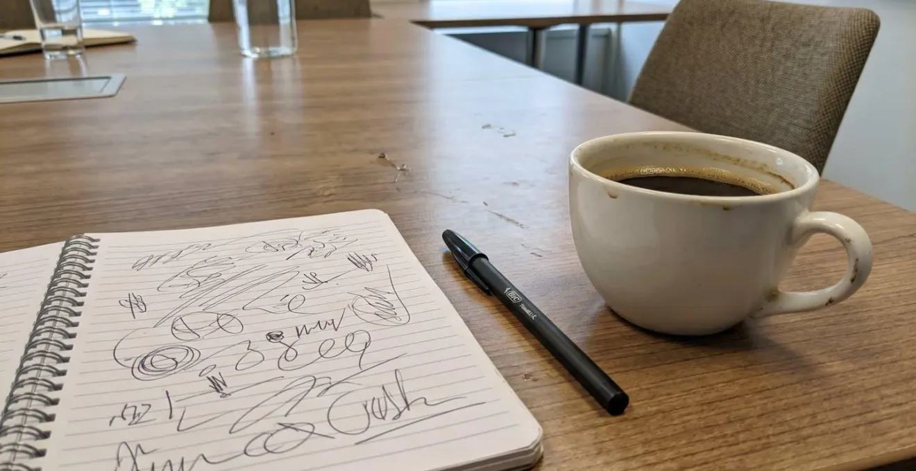 Gros plan sur une table de conférence contemporaine avec carnet ouvert, stylo de qualité et tasse de café sur surface en bois chaleureux, lumière naturelle douce